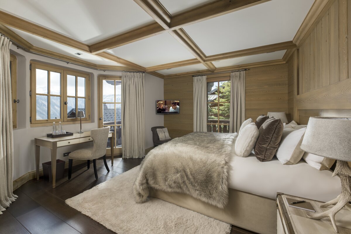 A cozy, wood-paneled chalet bedroom with a large bed, faux fur throw, writing desk by windows, and beamed ceiling, offering a warm mountain retreat.