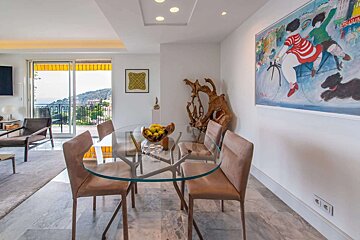 Bright, modern dining room with a glass table, four upholstered chairs, unique wall art, and a balcony with a stunning view of the sea and hills.