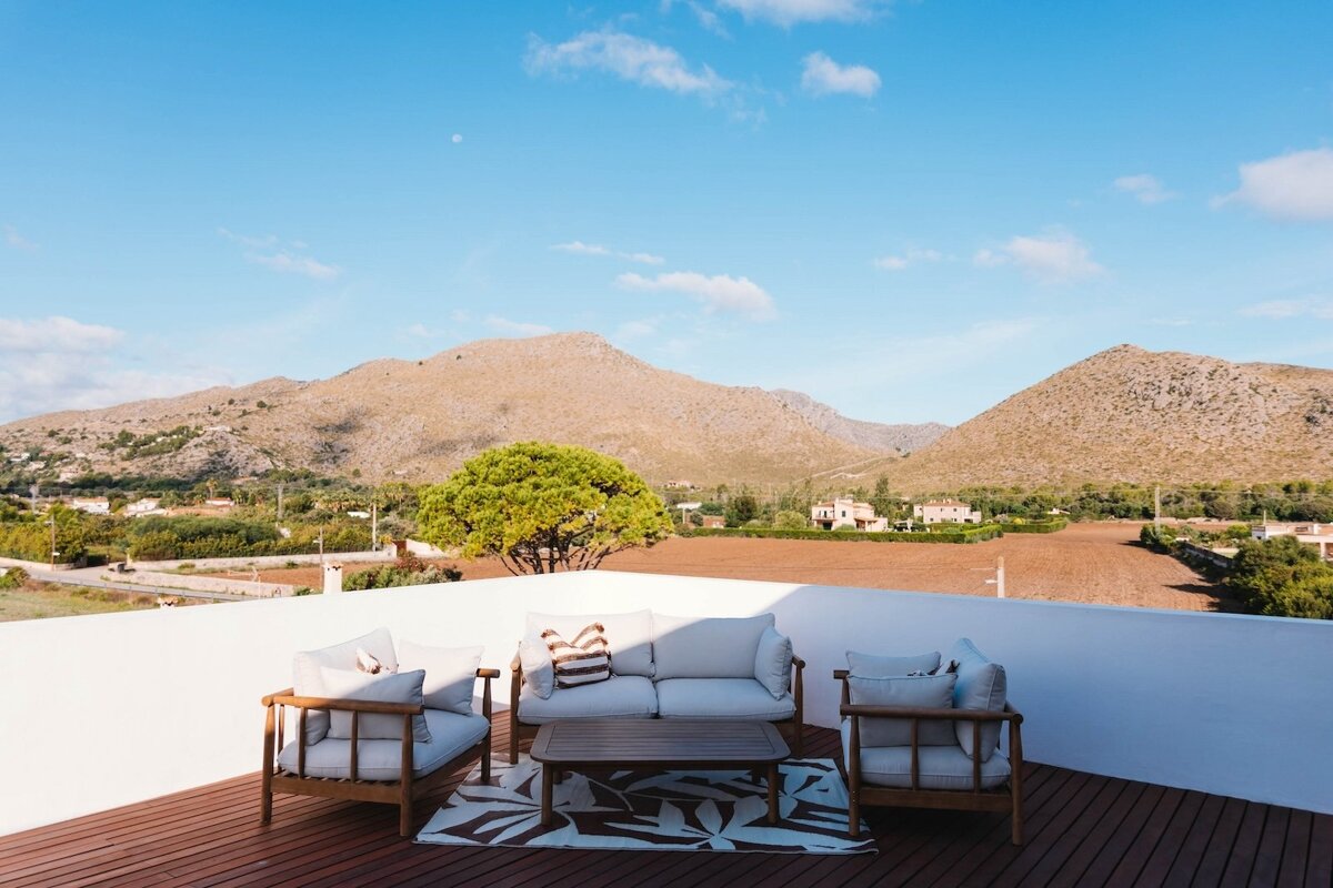 A patio with outdoor furniture offers scenic views of distant mountains, a green valley with scattered houses, under a bright blue sky with a faint moon.