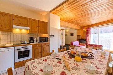 Cozy wooden chalet kitchen/dining area with a breakfast set on a patterned tablecloth, modern appliances, and a living space beyond.
