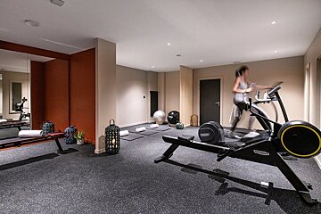A contemporary gym featuring a woman on a treadmill, a rowing machine, yoga mats, and stability balls. Modern design with dark flooring.