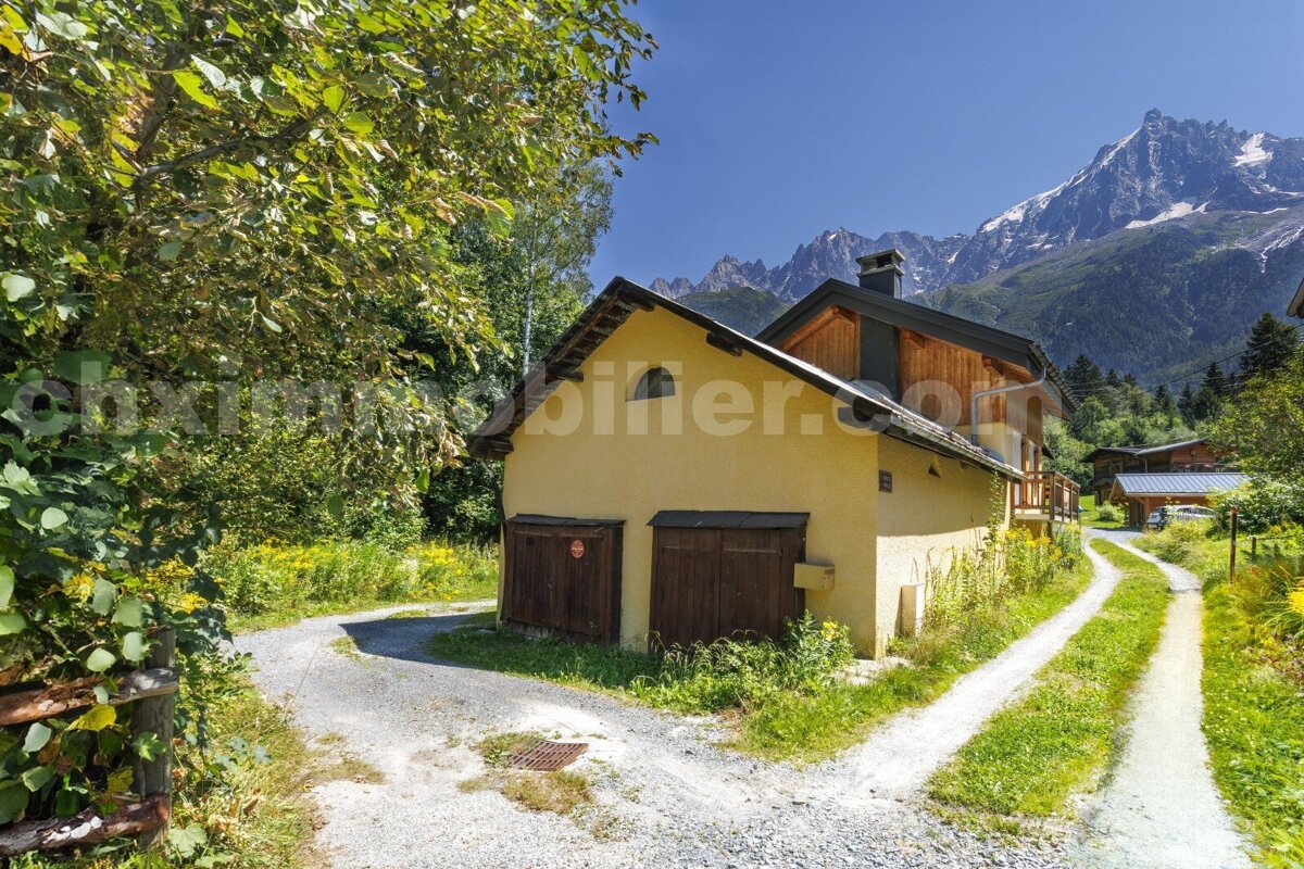 A yellow house with wooden doors stands by a gravel path, surrounded by green trees and wildflowers, with majestic mountains under a clear blue sky.