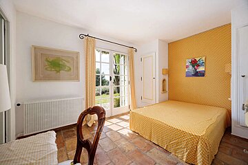 Bright yellow and white bedroom with a patterned bed, terracotta floor, and French doors opening to a sunny garden.