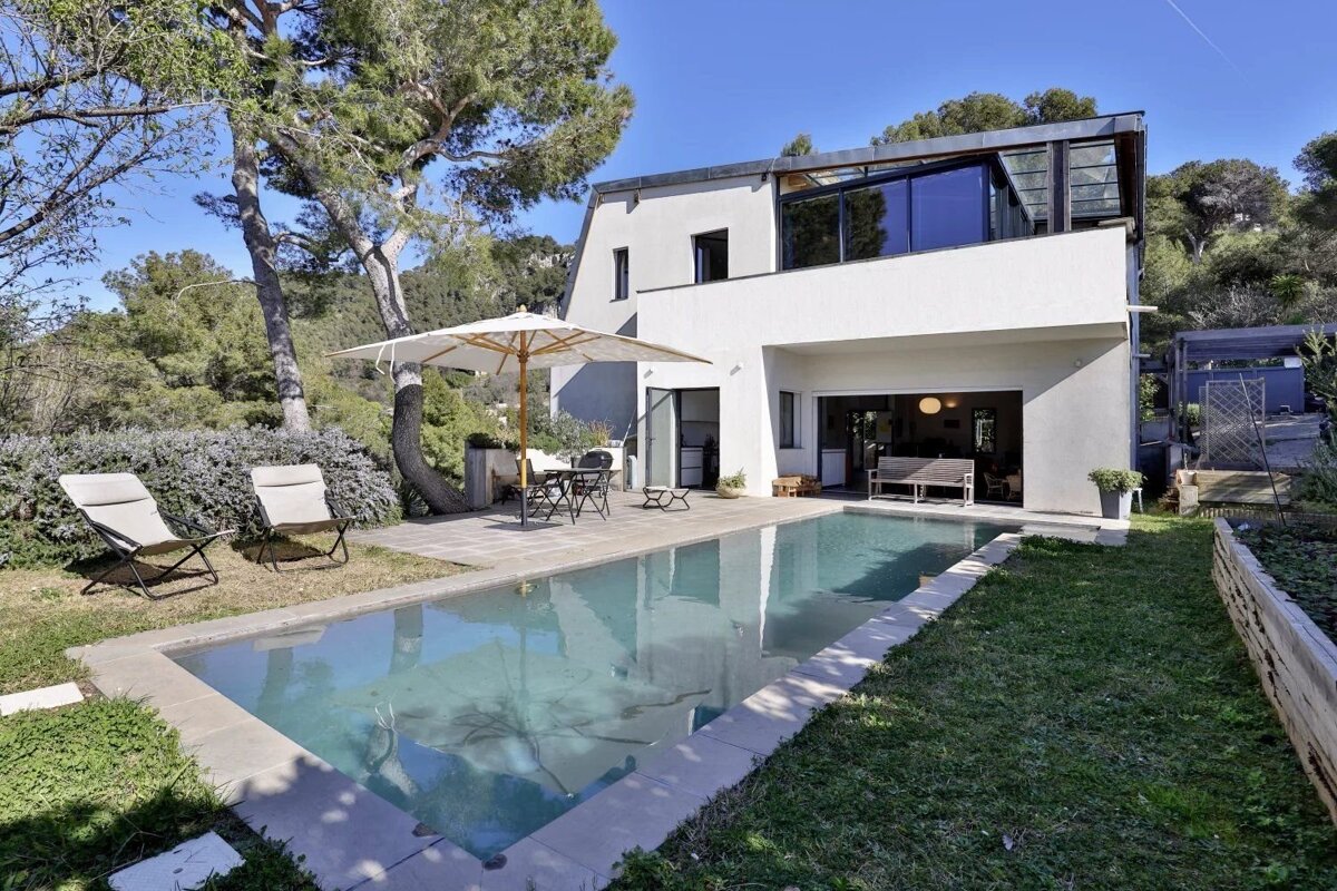 A modern white house with a swimming pool, patio, and garden, surrounded by lush trees under a clear blue sky.