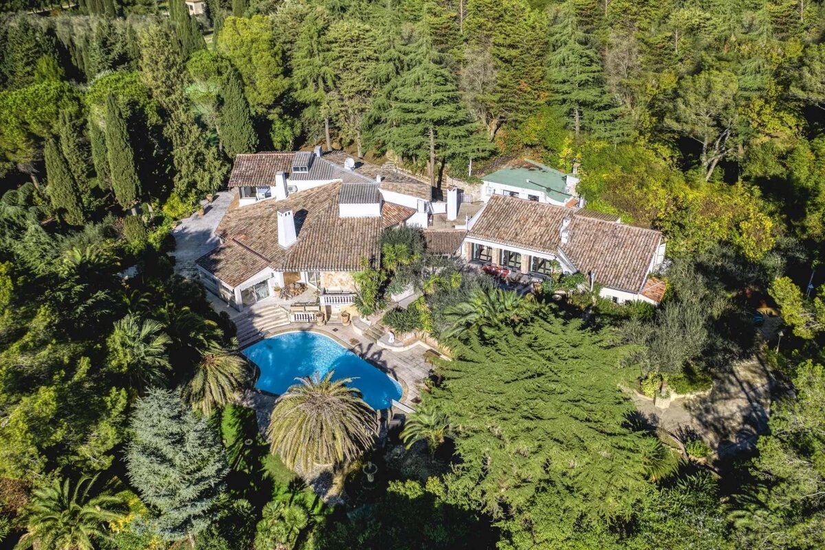 Aerial view of a sprawling villa with a swimming pool, completely surrounded by dense green forest and lush gardens.