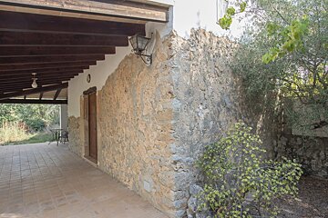 A rustic stone building with a covered tiled patio, an old-fashioned lantern, and lush green trees and shrubs under natural light.