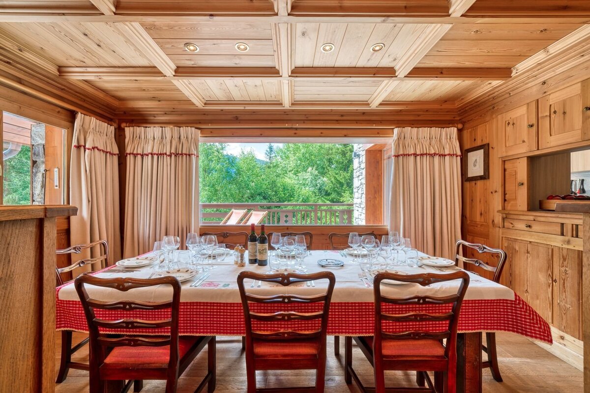 Warm, wood-paneled dining room with a large table set with a red checkered cloth, looking out to a green mountain forest view.