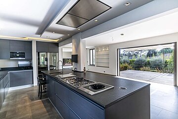 Modern open-plan kitchen with dark finishes, large island, and glass doors opening to a lush outdoor patio and garden.
