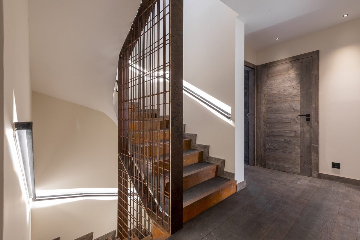 Wooden staircase with a rusted metal grid screen next to a rustic wooden door in a modern hallway with dark wood flooring.