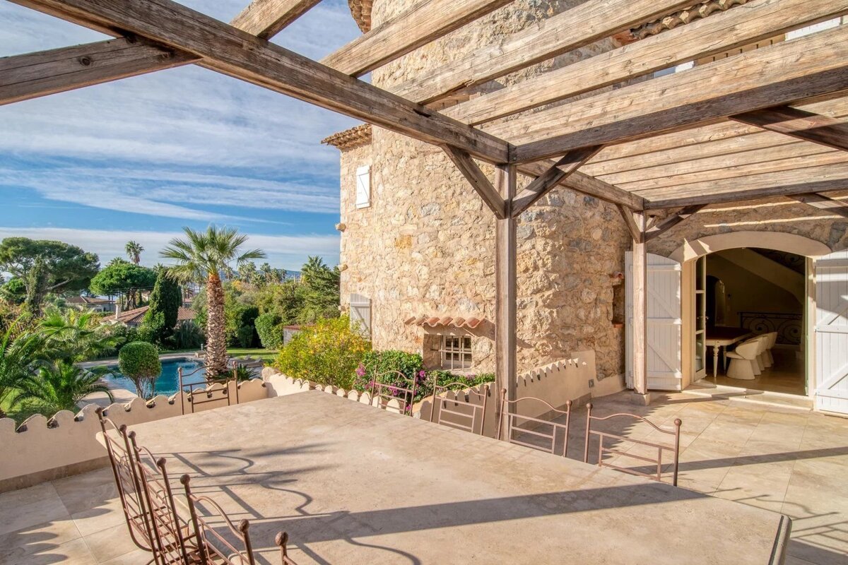 Stone villa with a pergola-covered patio, outdoor dining, lush garden, pool, and distant sea view under a blue sky.