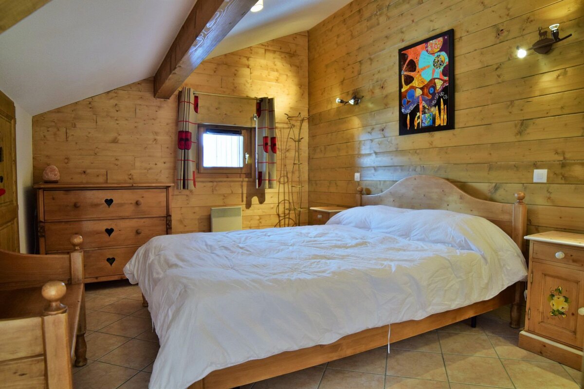 Rustic wooden bedroom featuring a large bed, heart-handled dresser, small window, and a vibrant abstract painting.