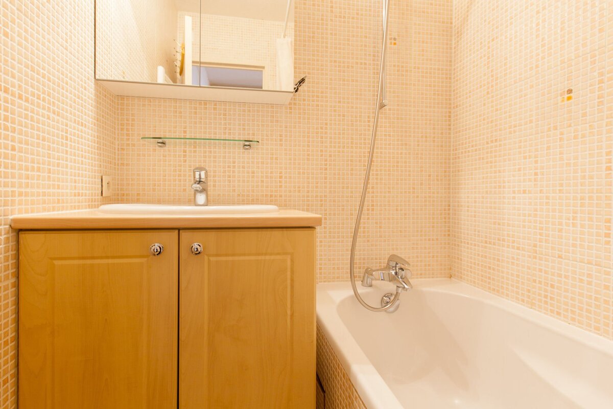 A small bathroom with beige mosaic tiles, a wooden vanity with a sink, a mirror cabinet, and a white bathtub with a shower attachment.