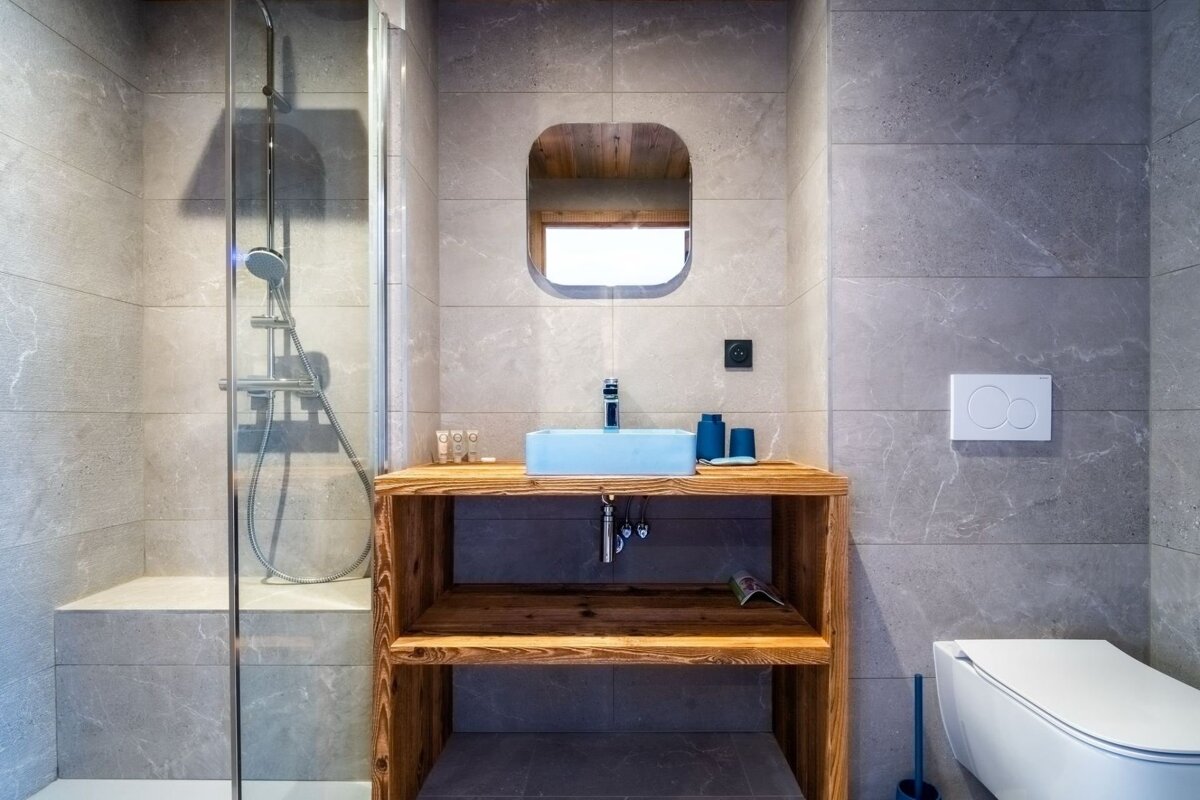 Modern bathroom with grey tiles, a glass shower, a rustic wooden vanity with a blue sink, and a wall-mounted toilet.