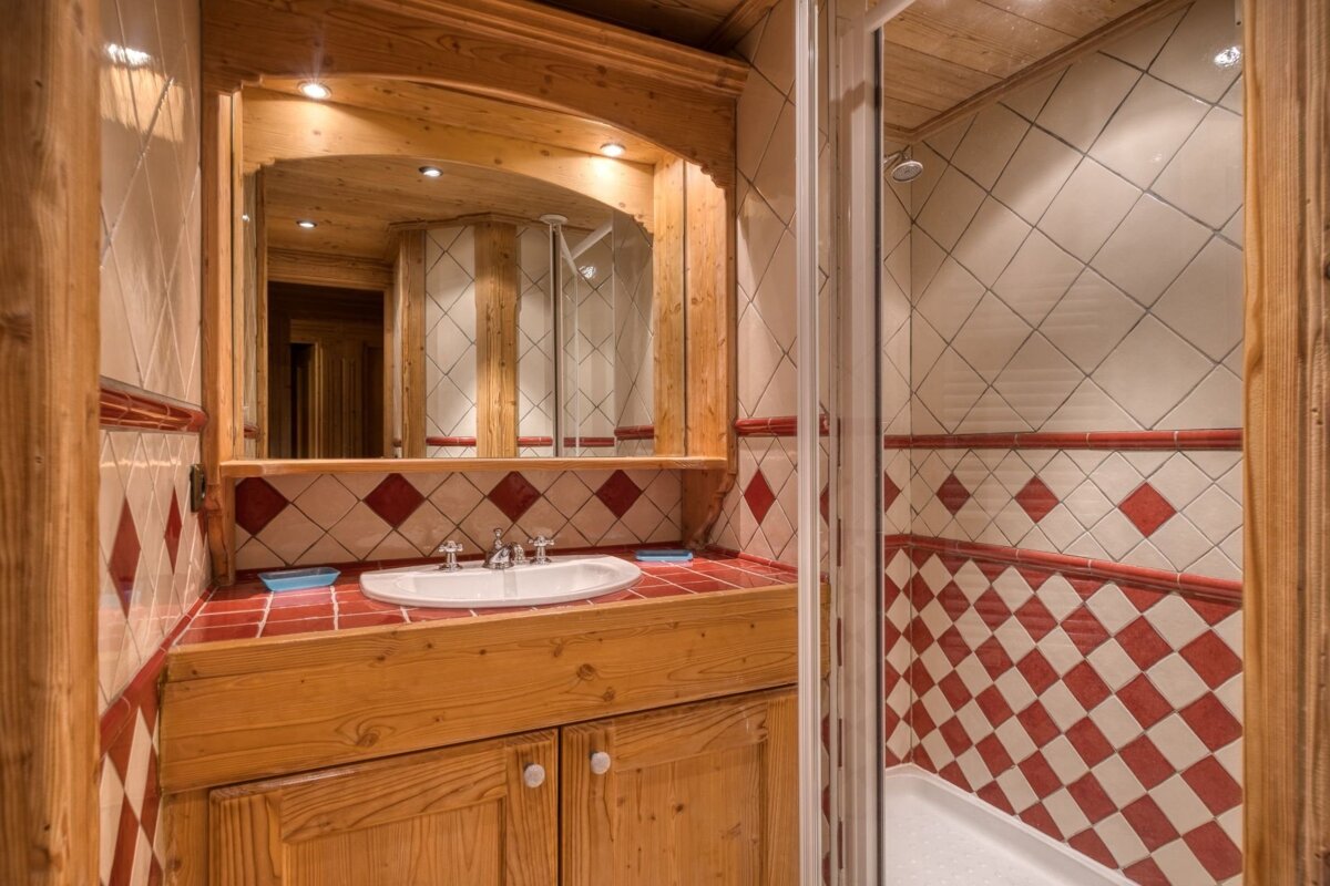 A cozy, rustic bathroom with a wooden vanity, white sink, and red and white diamond-patterned tiles. A shower is on the right.