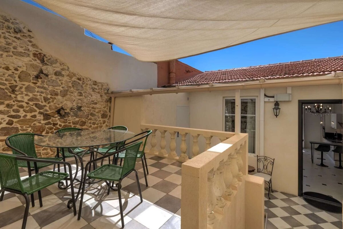 A sunny patio features a checkered floor, green dining set, stone wall, and cream balustrade under a sunshade. A room with a chandelier is visible.