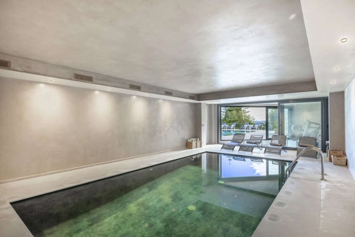 A modern indoor swimming pool with concrete walls and floor, featuring large glass sliding doors that open to an outdoor pool and lounge area.