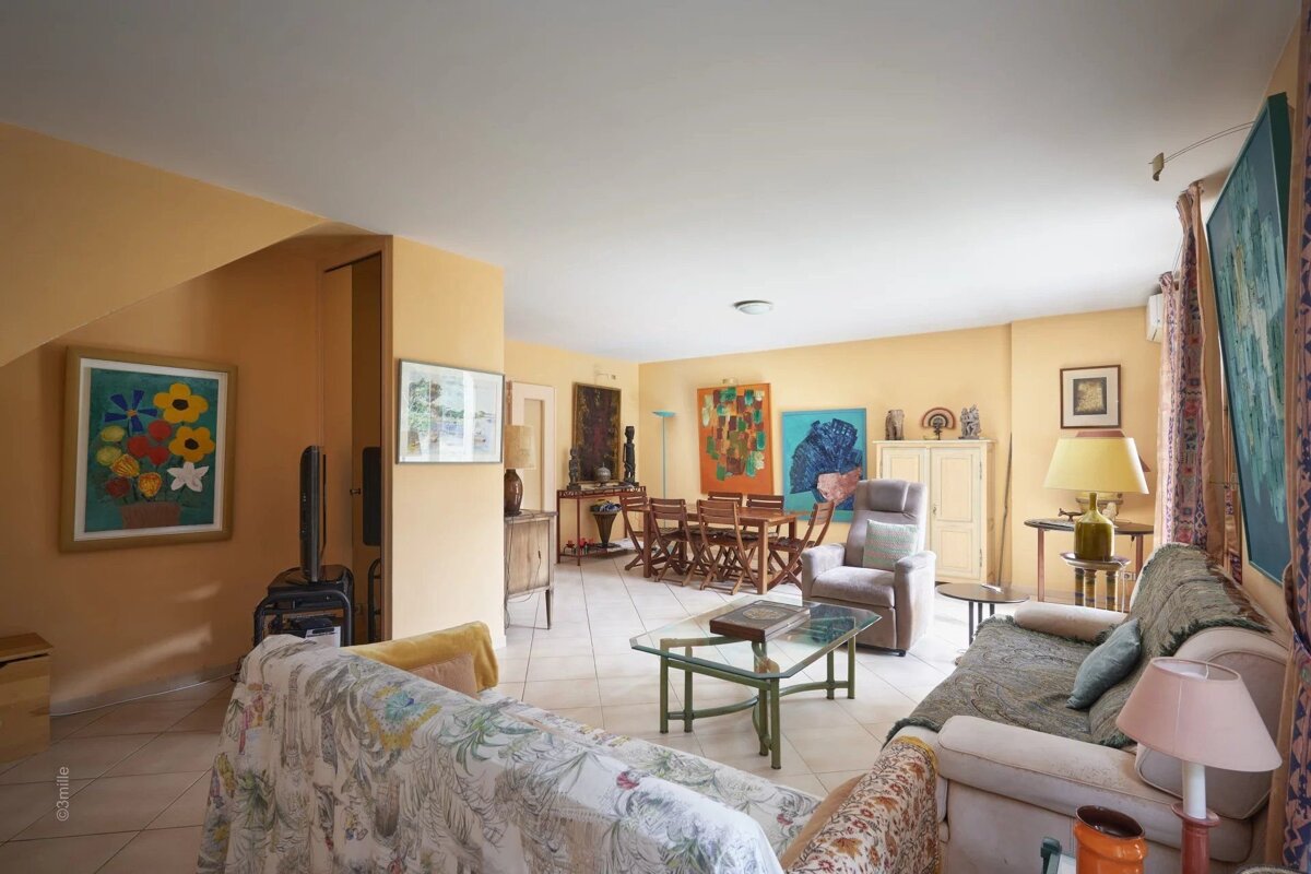 Eclectically decorated living room featuring warm yellow walls, colorful artwork, sofas, and a dining area in the background.