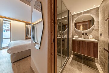 A modern bedroom with light wood accents and a mirror, adjacent to a chic bathroom with a vanity, illuminated oval mirror, and glass shower.
