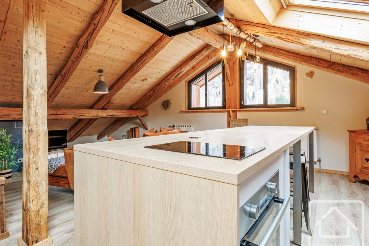 A rustic-modern kitchen with a large island and induction cooktop under a vaulted wooden ceiling with exposed beams and angular windows.