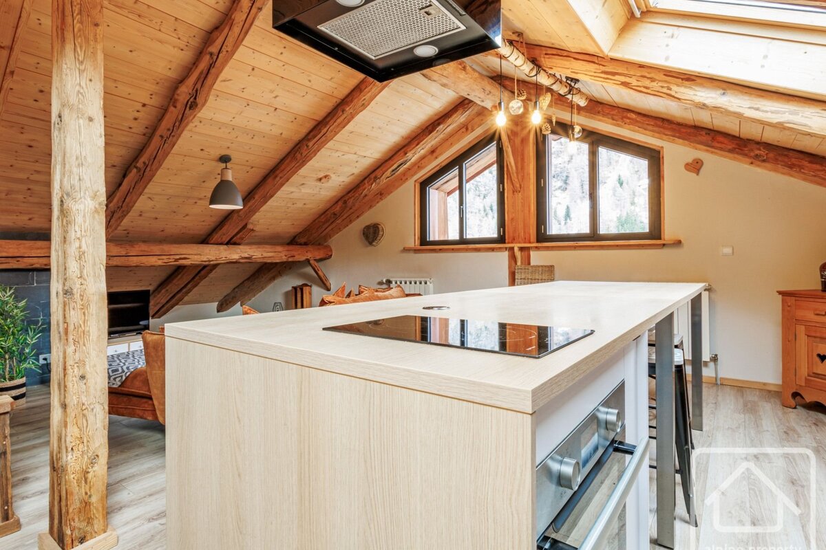 Rustic attic kitchen with exposed wooden beams, a modern island with a hob, and bright windows, creating a cozy, functional space.