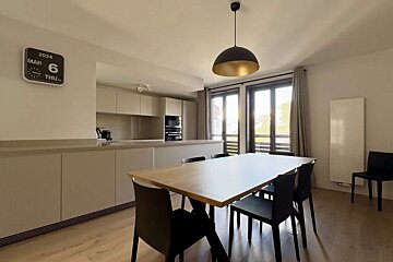 A modern open-plan kitchen and dining room with light wood floors, light cabinets, black chairs, and sunny windows. A calendar clock reads MAR 6 THU 2024.