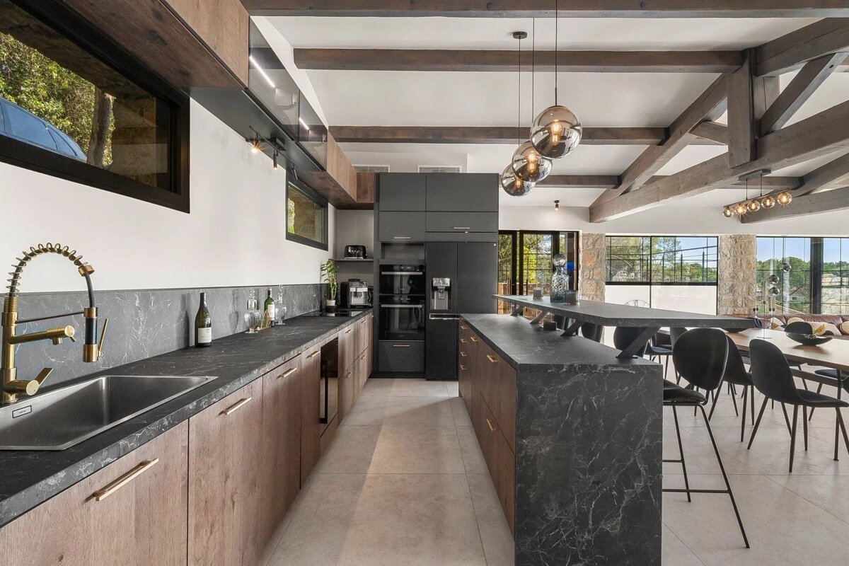 A modern open-concept kitchen with dark marble-look countertops, wood cabinetry, and a kitchen island with bar seating. Exposed wooden beams overhead.