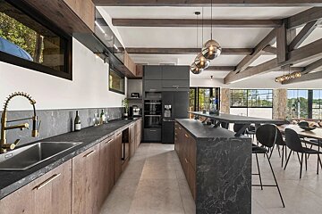 An expansive modern kitchen and dining space with dark marble counters, wood cabinetry, a large island, exposed ceiling beams, and big windows.