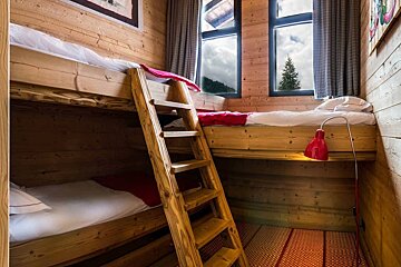 Cozy wooden cabin room featuring rustic bunk beds, a ladder, red bedding, and windows looking out to a pine tree.