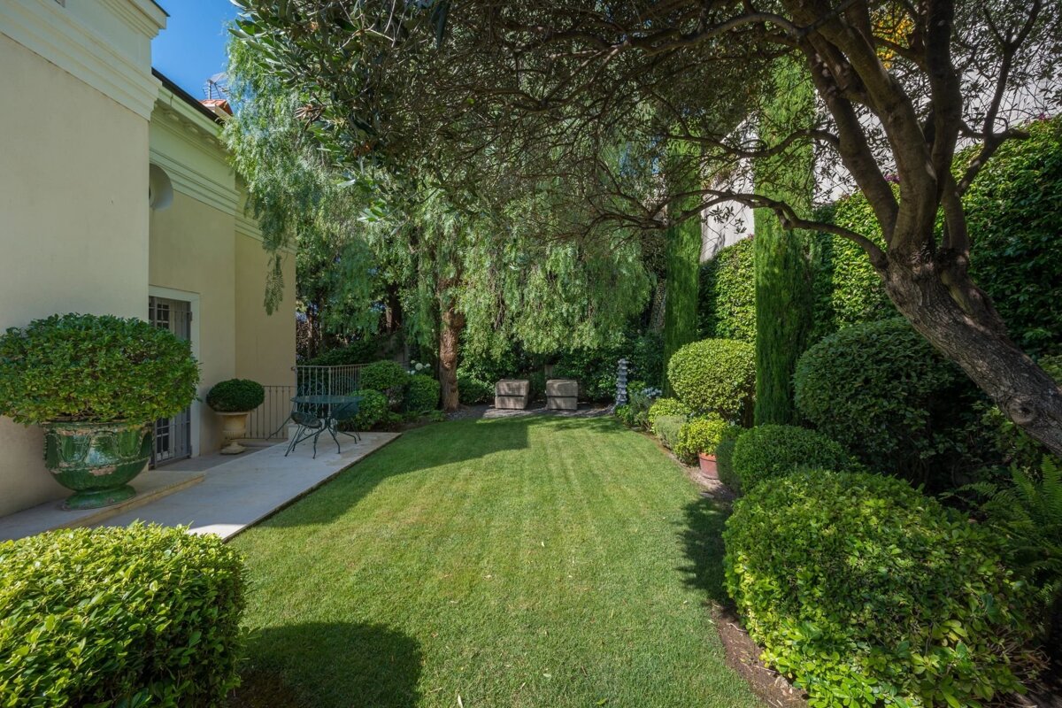 A lush, manicured garden with a green lawn, sculpted bushes, and mature trees next to a light-colored house.