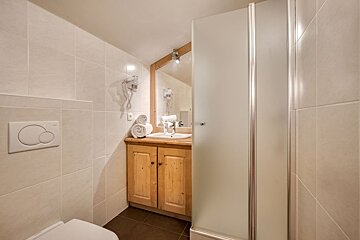 A small, light-tiled bathroom with a toilet, wooden vanity under a mirror, hairdryer, and a frosted glass shower cabin.