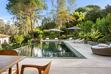 A luxurious infinity pool nestled in a vibrant green garden with lounge furniture and umbrellas, all under a bright sky.