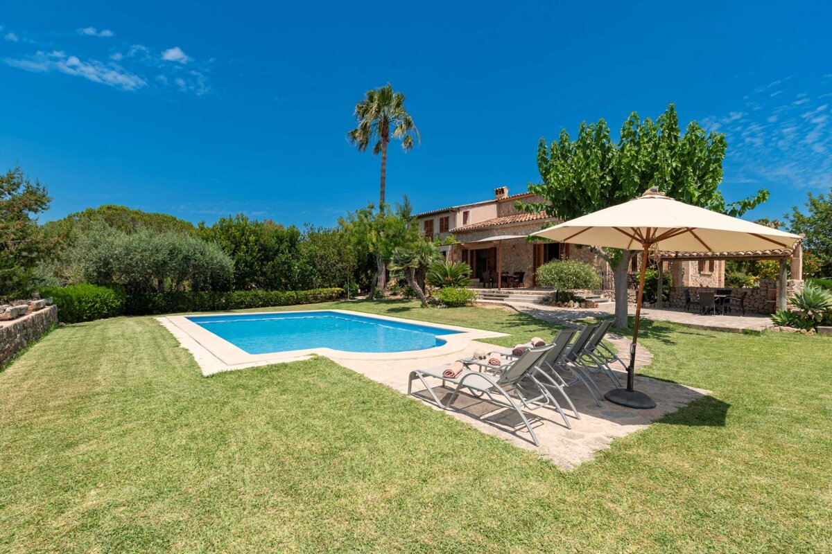 Sunny villa with a sparkling blue pool, green lawn, and sun loungers under a bright blue sky.