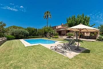Sunny villa with a sparkling blue pool, green lawn, and sun loungers under a bright blue sky.