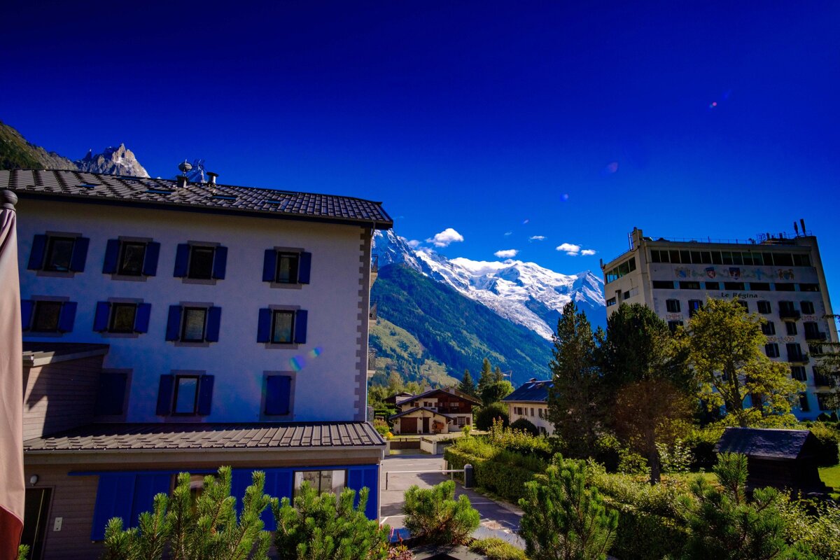 Bright blue sky frames buildings with blue shutters and a hotel, backed by majestic snow-capped mountains and green valleys.