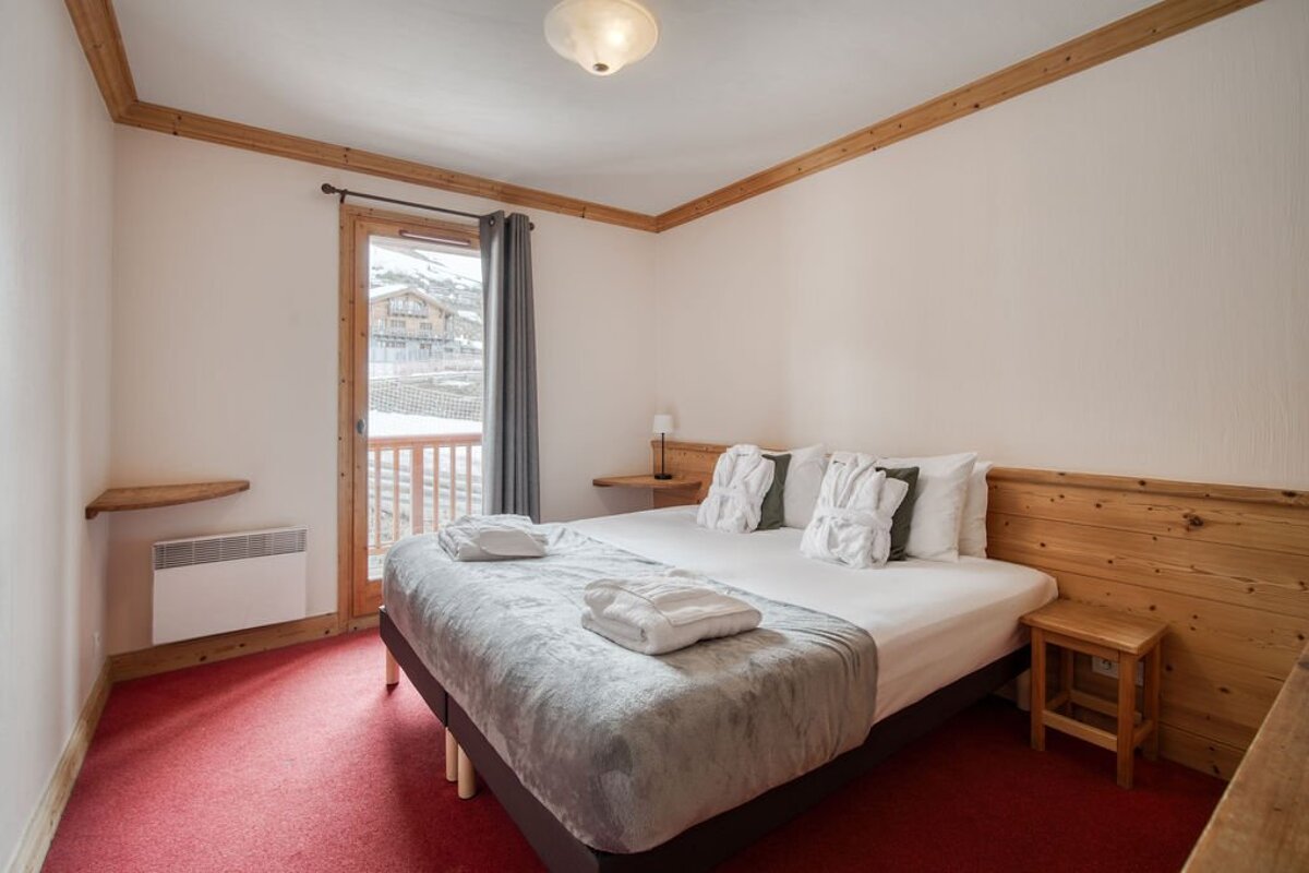 Alpine style bedroom with a large bed, red carpet, wooden details, and a balcony offering a view of snowy mountains and a village.