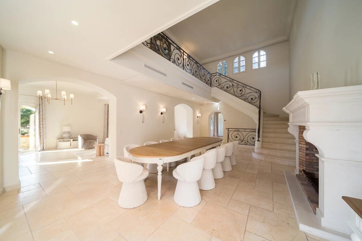 Spacious, elegant room with a long dining table, white chairs, a grand staircase with ornate railing, a large fireplace, and light tiled floors.