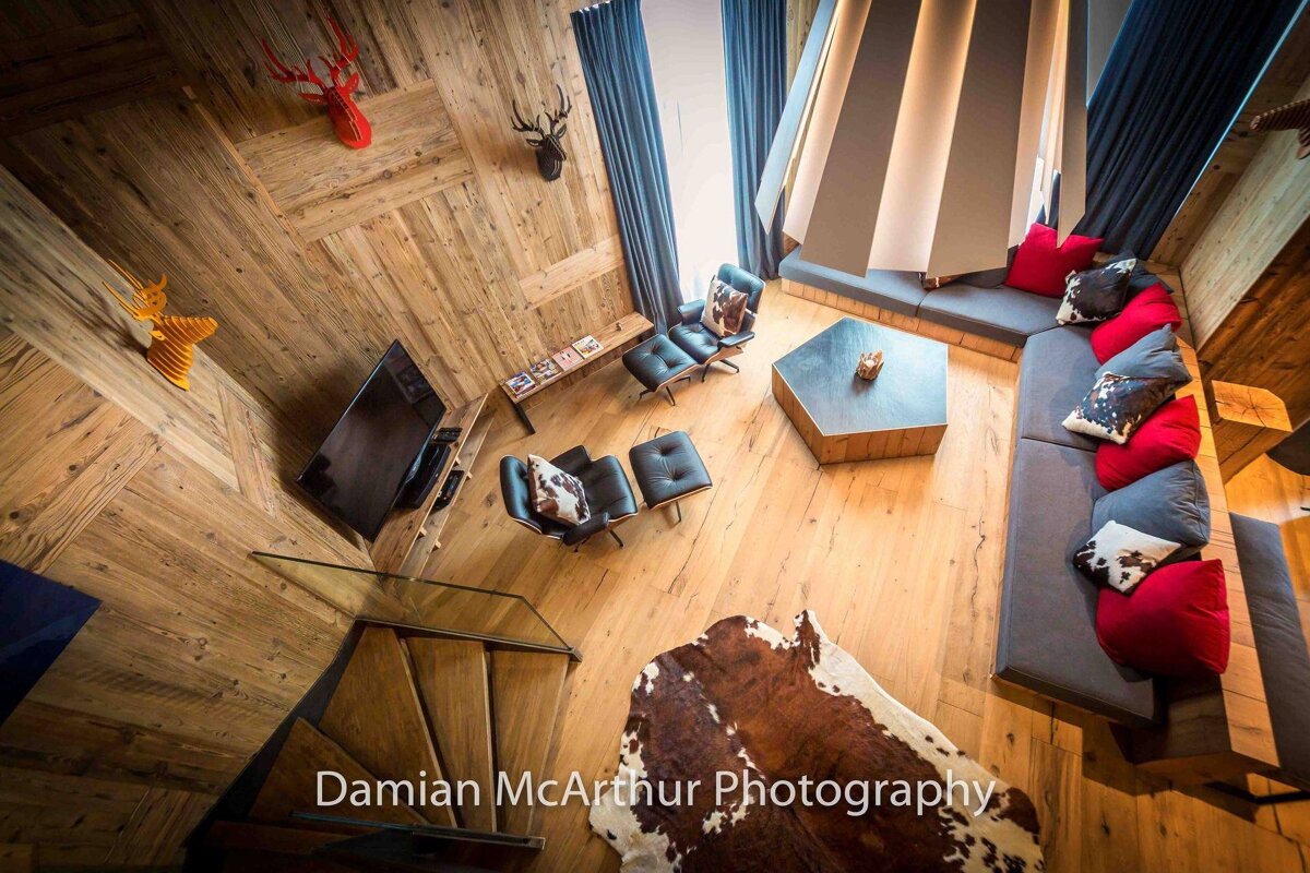 High-angle view of a modern living room with extensive wood paneling, a large sectional sofa with red and cowhide pillows, chic chairs, and deer head decor.