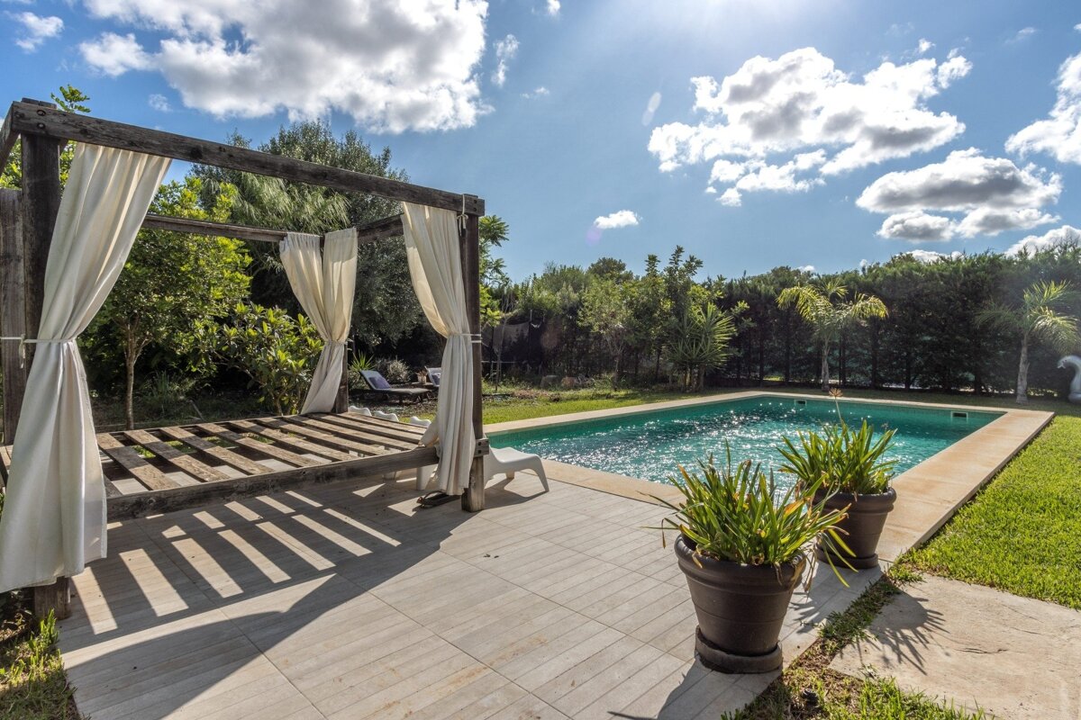 Sunny poolside scene with a sparkling blue pool, a wooden pergola with white curtains, lush green landscaping, and potted plants under a partly cloudy sky.
