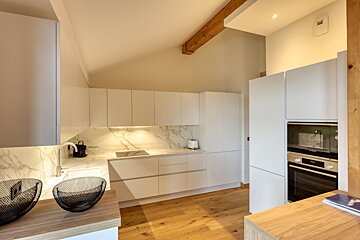 A sleek, modern kitchen with white handleless cabinets, marble backsplash, light wood floors, and a striking beamed, sloped ceiling.