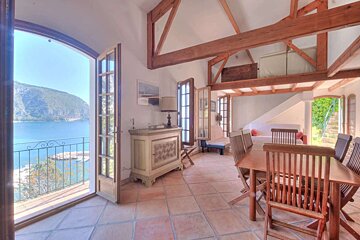 Rustic open living room with terracotta tiles, wood beams, and a dining area. French doors lead to a balcony with a stunning blue sea and mountain view.