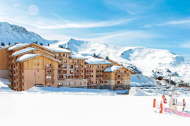 A snowy mountain resort features large chalet-style buildings under a bright blue sky, with white peaks and ski slopes in the background.