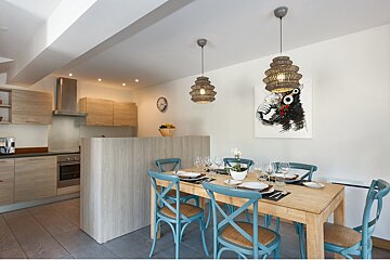 A bright, open-plan kitchen and dining room. Features light wood cabinetry, a set table with blue chairs, and a quirky monkey painting.