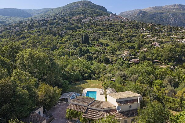 Aerial view: a house with a pool in a green valley, backed by tree-covered mountains and a village on a distant peak.