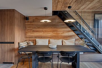 A contemporary dining area with a dark wooden table, built-in bench, warm wood paneling, and a sleek black metal & glass staircase.