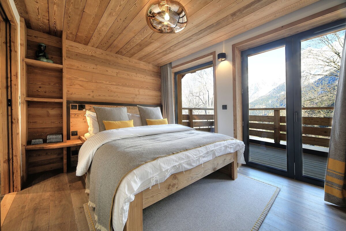 A cozy, rustic bedroom featuring wood-paneled walls, a large bed with gray and yellow accents, and a balcony with stunning mountain views.