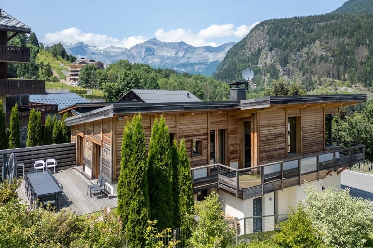 Modern two-story wooden chalet with a large deck, nestled in a lush mountain valley with rocky peaks behind.
