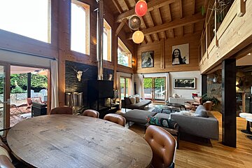 Spacious wooden chalet interior with high ceilings, large windows, a dining area, and a cozy living room featuring a fireplace and modern decor.