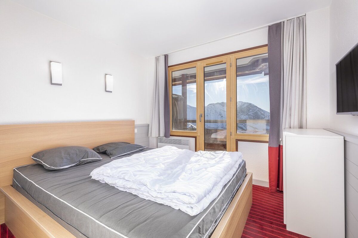 A cozy bedroom with a wooden bed, grey bedding, and white duvet. A large window reveals snowy mountain views. Red striped carpet and a wall-mounted TV.