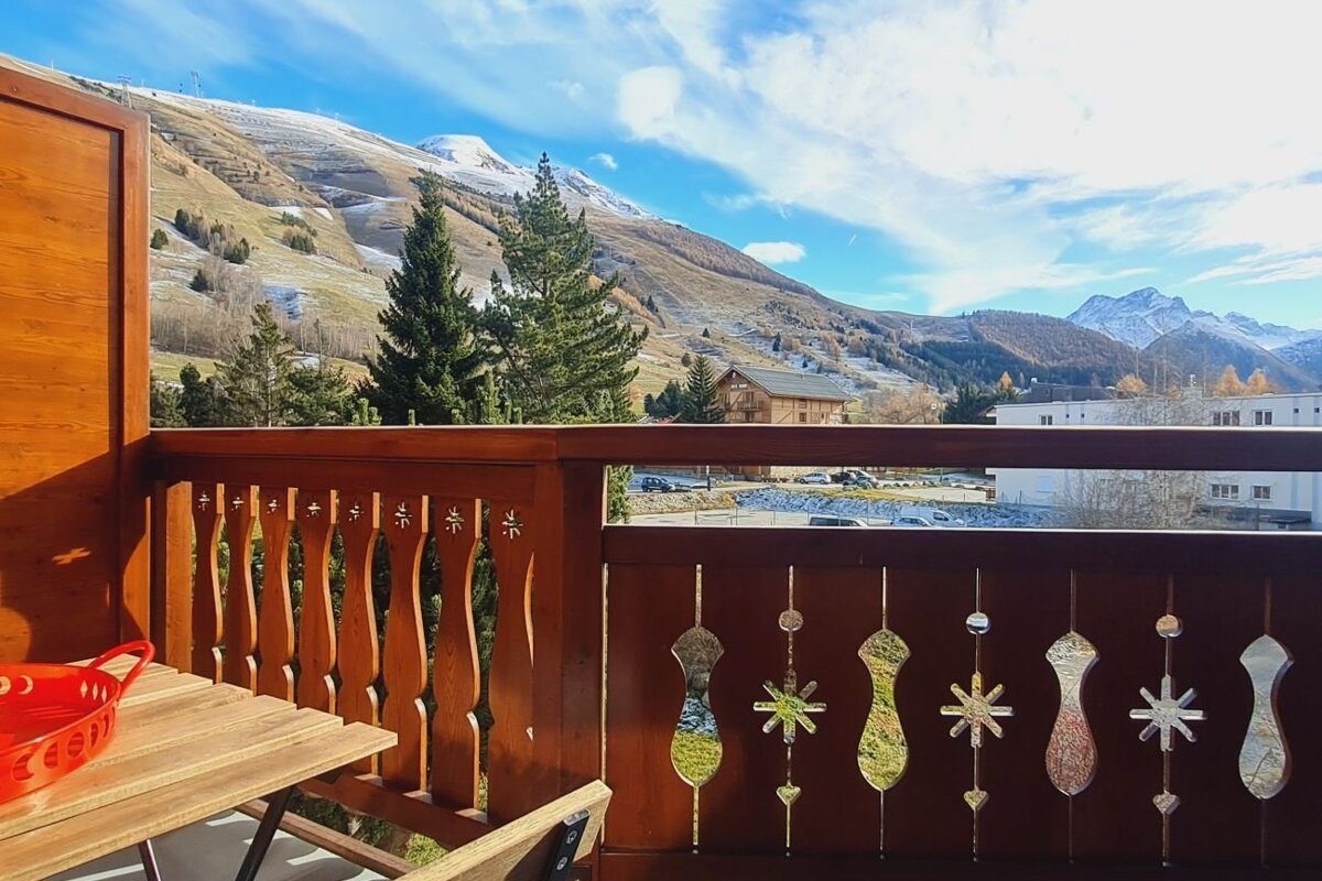 Wooden balcony overlooking a picturesque mountain landscape with evergreen trees and snow-capped peaks under a blue sky.
