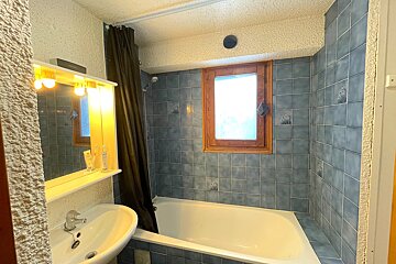 A blue-tiled bathroom features a white bathtub with a dark shower curtain, a sink with an illuminated mirror, and a small wooden window.